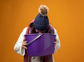 Free photo young ill woman wearing robe winter hat and scarf having nausea holding plastic bucket vomiting into it isolated on orange wall