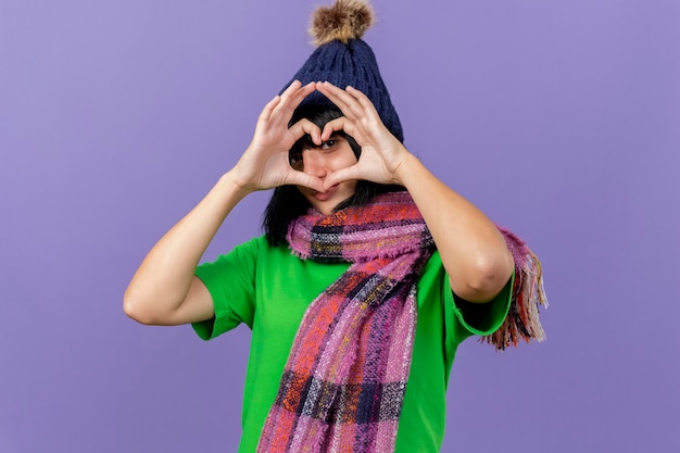Молодая больная женщина в зимней шапке и шарфе делает знак сердца, глядя вперед сквозь него, изолированную на фиолетовой стене с копией пространства