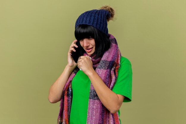 Young ill woman wearing winter hat and scarf talking on phone looking straight holding napkin coughing isolated on olive green wall with copy space