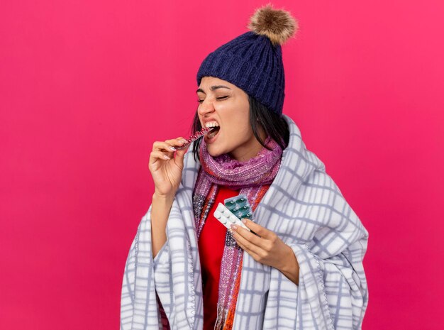 Young ill woman wearing winter hat and scarf wrapped in plaid holding packs of pills biting one of them with closed eyes isolated on pink wall with copy space