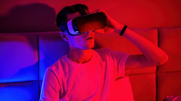 Молодой впечатленный человек в очках виртуальной реальности с синей и красной подсветкой в комнате на кровати. Развлечения дома