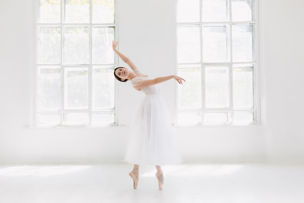 젊고 믿을 수 없을만큼 아름다운 발레리나는 흰색 스튜디오에서 포즈를 취하고 춤을 추고 있습니다.