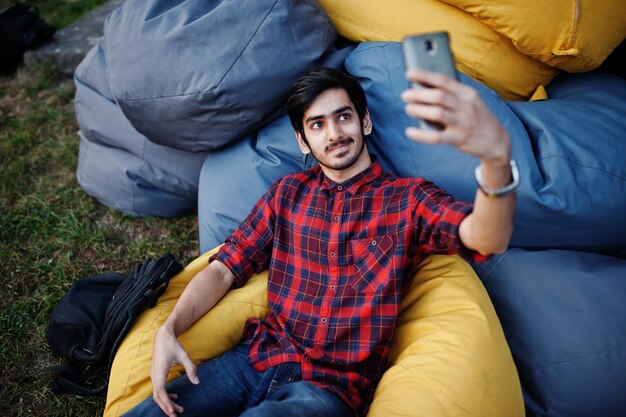 체크 무늬 셔츠와 청바지를 입은 젊은 인도 학생 남자가 야외 베개에 앉아 휴식을 취하고 셀카를 만듭니다.