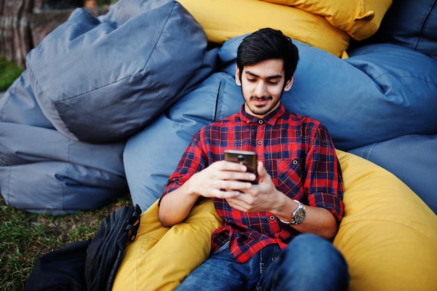 체크 무늬 셔츠와 청바지를 입은 젊은 인도 학생 남자가 야외 베개에 앉아 휴식을 취하며 휴대폰으로 시간을 보낸다