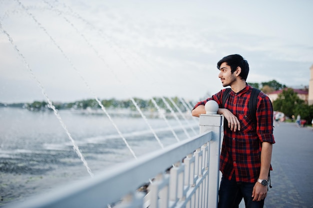 Young indian student man at checkered shirt and jeans with backpack posed on evening city against fountains