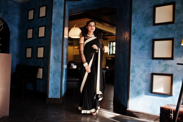 若いインド人女性は、フレームの壁に対してレストランでポーズをとったエレガントな黒のサリーで着用します