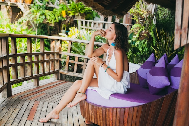 Young innocent pure beautiful woman dreaming, sitting at couch in white dress, romantic, lyrical, thinking, green tropical nature, summer, relaxed, chilling, legs, resort hotel