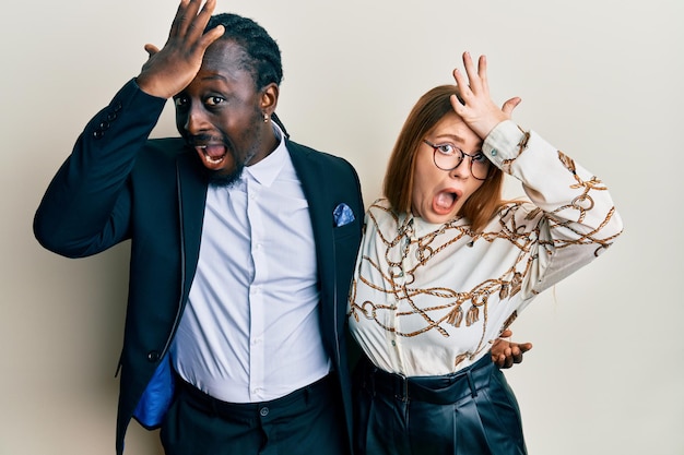 Young interracial couple wearing business and elegant clothes surprised with hand on head for mistake, remember error. forgot, bad memory concept.