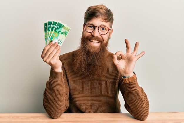 20 イスラエル シェケル紙幣を保持している若いアイルランドの赤毛の男は、テーブルの上に座って指で ok サインをし、フレンドリーなジェスチャーの優れたシンボルに笑みを浮かべて