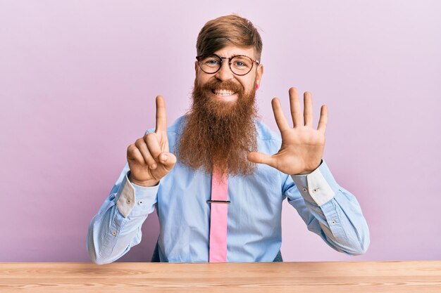 Молодой ирландский рыжий мужчина в деловой рубашке и галстуке сидит на столе, показывая и указывая пальцами номер шесть, улыбаясь уверенно и счастливо.