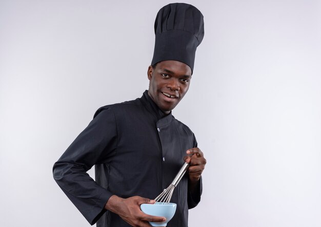 Young joyful afro-american cook in chef uniform holds bowl and whisk on white  with copy space