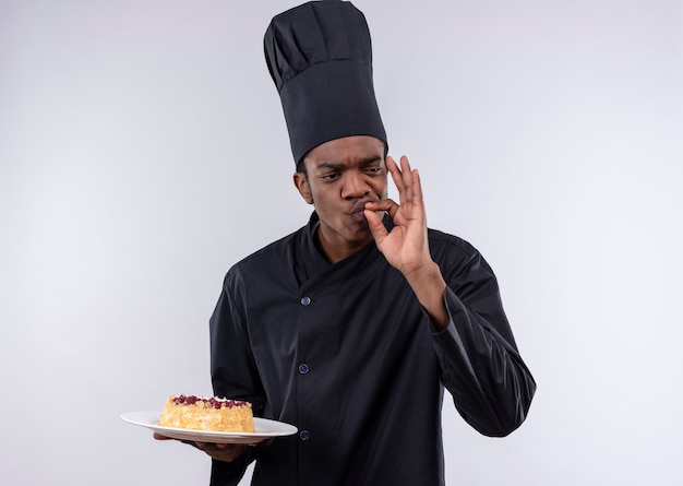 Free photo young joyful afro-american cook in chef uniform holds cake on plate and gestures tasty delicious isolated on white wall