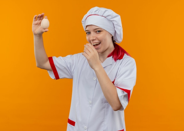 Young joyful blonde female chef in chef uniform holds eggs isolated on orange wall