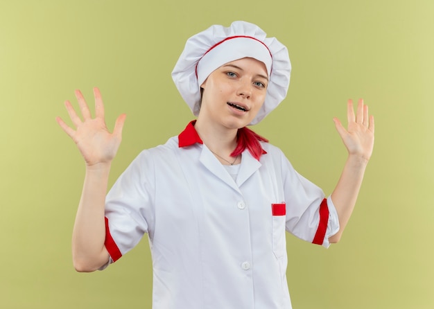 Il giovane chef femminile biondo gioioso in uniforme del cuoco unico tiene le mani in alto isolato sulla parete verde