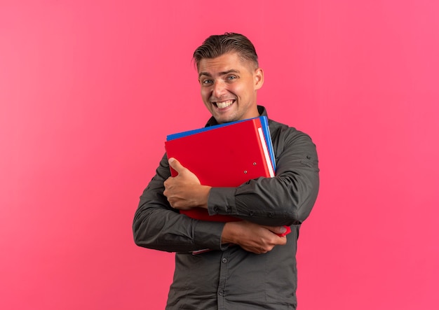 young joyful blonde handsome man holds file folders isolated on pink background with copy space
