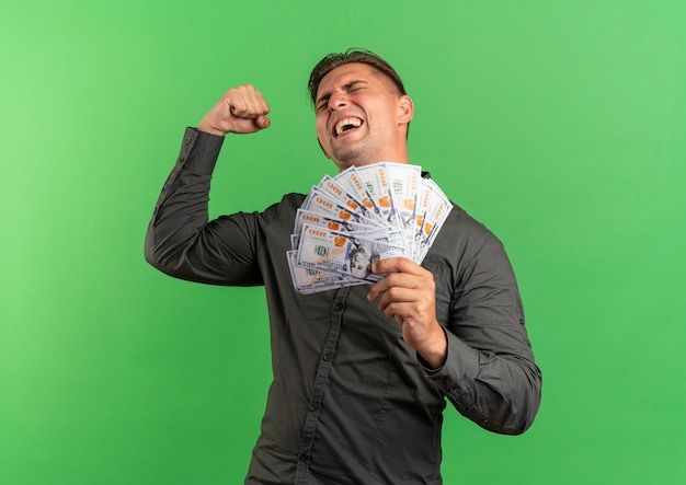 Young joyful blonde handsome man holds money with raised fist up isolated on green space with copy space