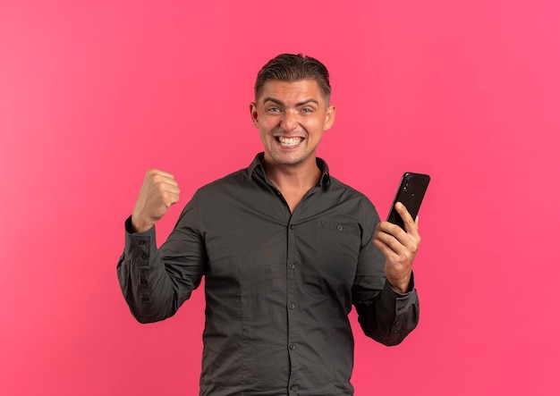 Young joyful blonde handsome man holds phone and keeps fist up looking at camera isolated on pink background with copy space