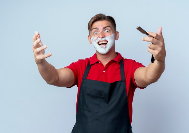 Young joyful blonde male barber in uniform smeared face with shaving foam holding straight razor isolated on white space with copy space