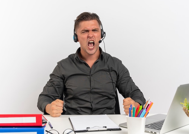 Free photo young joyful blonde office worker man on headphones sits and puts fists at desk with office tools using laptop isolated on white background with copy space