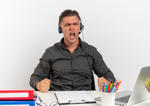 Молодой радостный белокурый человек офисного работника в наушниках сидит и кладет кулаки на стол с офисными инструментами, используя ноутбук, изолированный на белом фоне с копией пространства