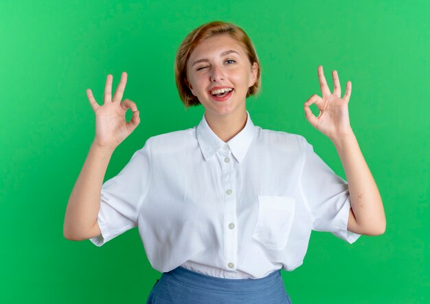 Young joyful blonde russian girl blinks eye gesturing ok sign with two hands isolated on green background with copy space