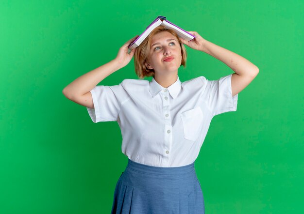 Foto gratuita la giovane ragazza russa bionda allegra tiene il libro sopra la testa isolata su fondo verde con lo spazio della copia