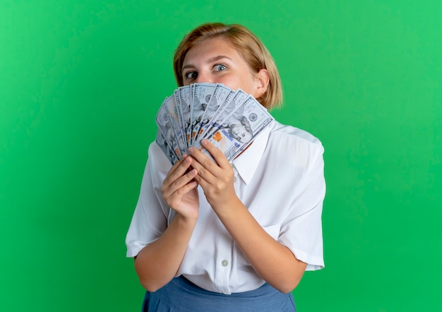 Free photo young joyful blonde russian girl holds and looks over money isolated on green background with copy space