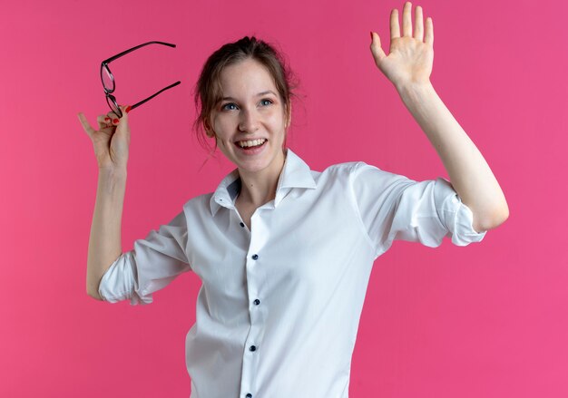 Young joyful blonde russian girl holds optical glasses and raises hand up looking forward on pink  with copy space