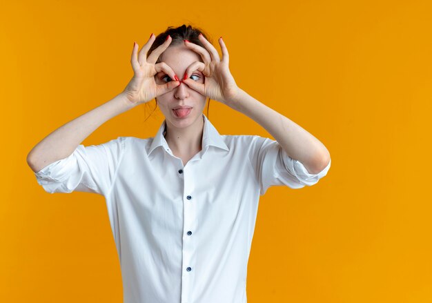 Young joyful blonde russian girl looks through fingers and stucks out tongue on orange  with copy space