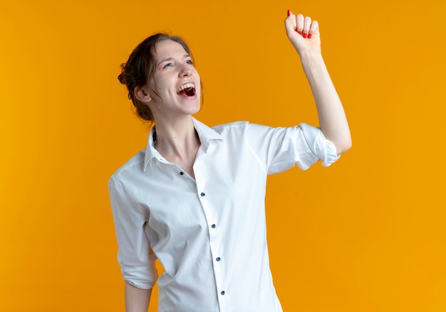 Young joyful blonde russian girl raises fist up looking at side isolated on orange space with copy space
