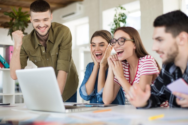Молодые радостные деловые люди счастливо работают на ноутбуке вместе Группа улыбающихся мужчин и женщин, проводящих время в современном уютном офисе