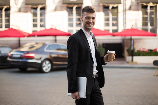 Young joyful businessman in black suit and white shirt with wireless earphones holding laptop and coffe in hands while happily looking in camera with beautiful street on background