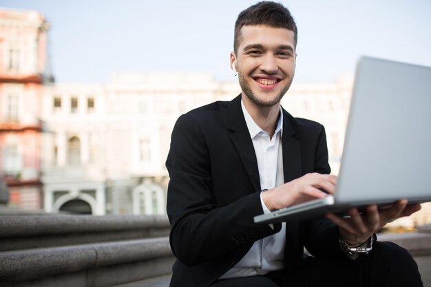 Young joyful businessman in classic black jacket and white shirt with wireless earphones happily looking in camera with laptop in hands outdoor