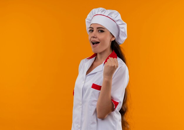 Young joyful caucasian cook girl in chef uniform holds fist up isolated on orange wall with copy space