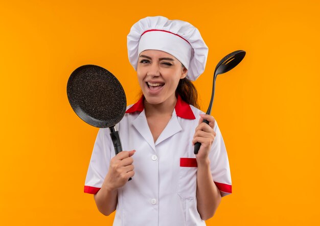 요리사 제복을 입은 젊은 즐거운 백인 요리사 소녀는 프라이팬을 보유하고 주걱은 복사 공간이있는 주황색 벽에 고립 된 눈을 깜박입니다.