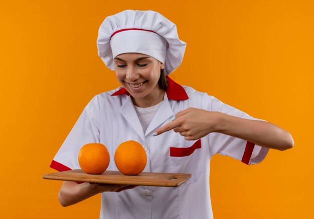 Young joyful caucasian cook girl in chef uniform holds and points at oranges on cutting board isolated on orange space with copy space