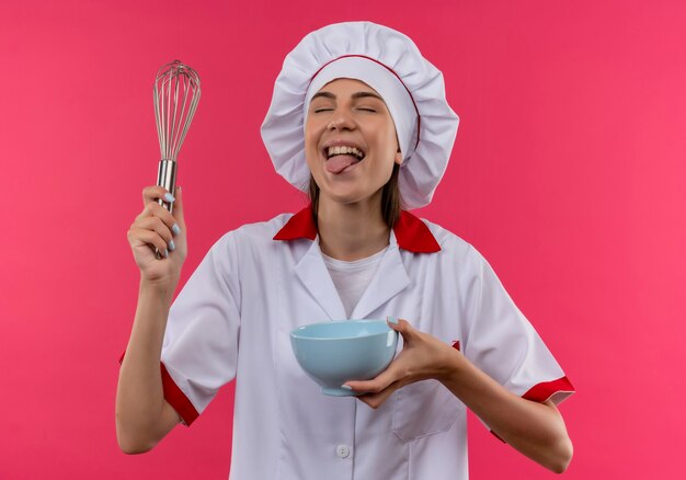 Young joyful caucasian cook girl in chef uniform holds whisk and bowl sticking tongue out on pink  with copy space