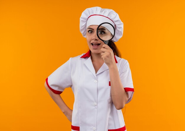 Молодая веселая кавказская девушка-повар в униформе шеф-повара смотрит через увеличительное стекло или лупу, изолированную на оранжевой стене с копией пространства