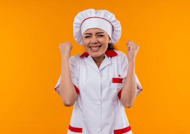 Молодая радостная кавказская девушка-повар в униформе шеф-повара поднимает кулаки вверх изолированной на оранжевой стене с копией пространства