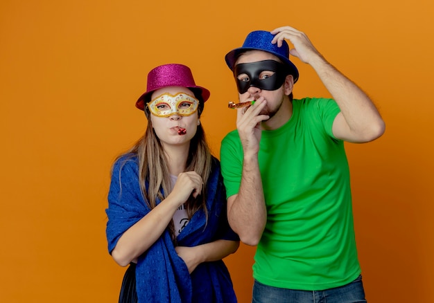 ピンクと青の帽子をかぶって、笛を吹いてオレンジ色の壁に孤立して見える仮面舞踏会のアイマスクを身に着けている若い楽しいカップル