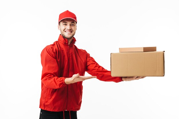 Young joyful delivery man in red cap and jacket happily looking in camera while giving package to customer over white background