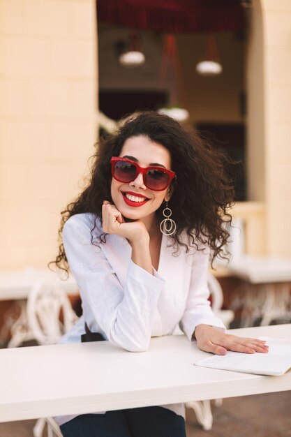 Young joyful lady with dark curly hair in sunglasses standing and happily looking in camera while spending time in summer terrace of cafe