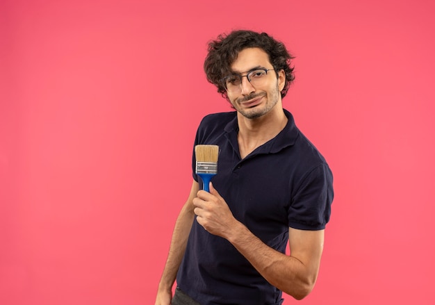 Young joyful man in black shirt with optical glasses holds brush and looks isolated on pink wall