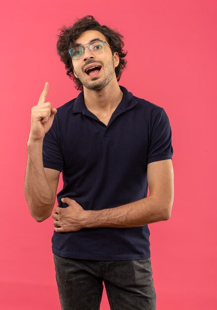 Young joyful man in black shirt with optical glasses points up isolated on pink wall
