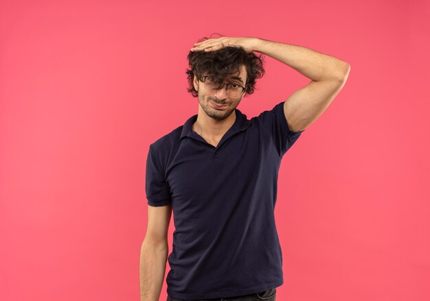 Young joyful man in black shirt with optical glasses puts hand on head and looks isolated on pink wall