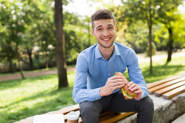 Young joyful man in blue shirt sitting on bench with sandwich in hands and cup of coffee to go near happily looking in camera while spending time in cozy park