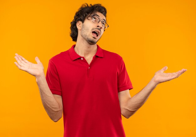 Young joyful man in red shirt with optical glasses holds hands open isolated on orange wall