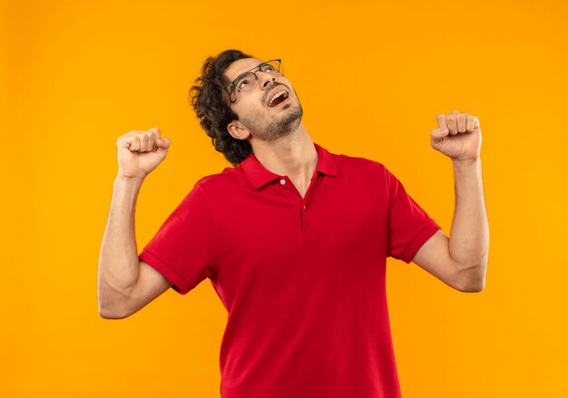 Young joyful man in red shirt with optical glasses raises fists up and looks up isolated on orange wall