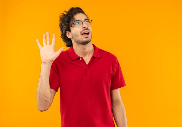 Young joyful man in red shirt with optical glasses raises hand and looks up isolated on orange wall