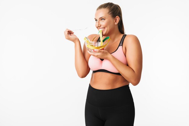 Young joyful plump woman in sporty top pretending that eat measuring tape from bowl while happily looking aside over white background
